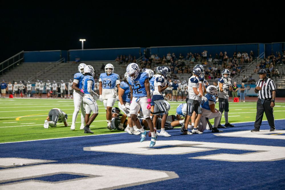 High school football players in blue and white uniforms celebrate on the field during a night game while a referee looks on.