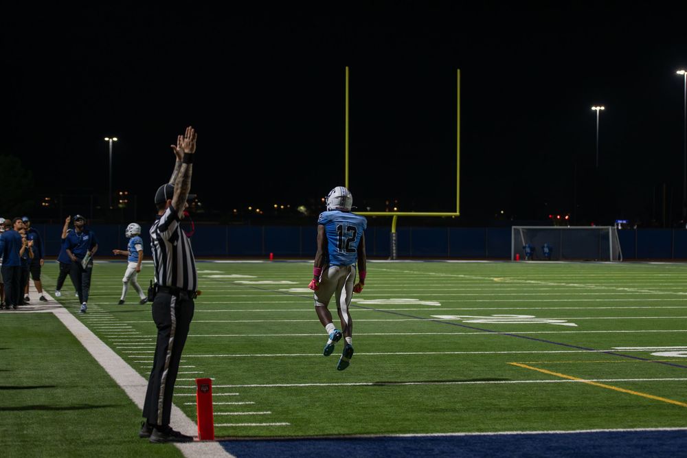 Football player in a light blue uniform runs downfield near the goalpost during a night game while a referee raises one arm to signal.