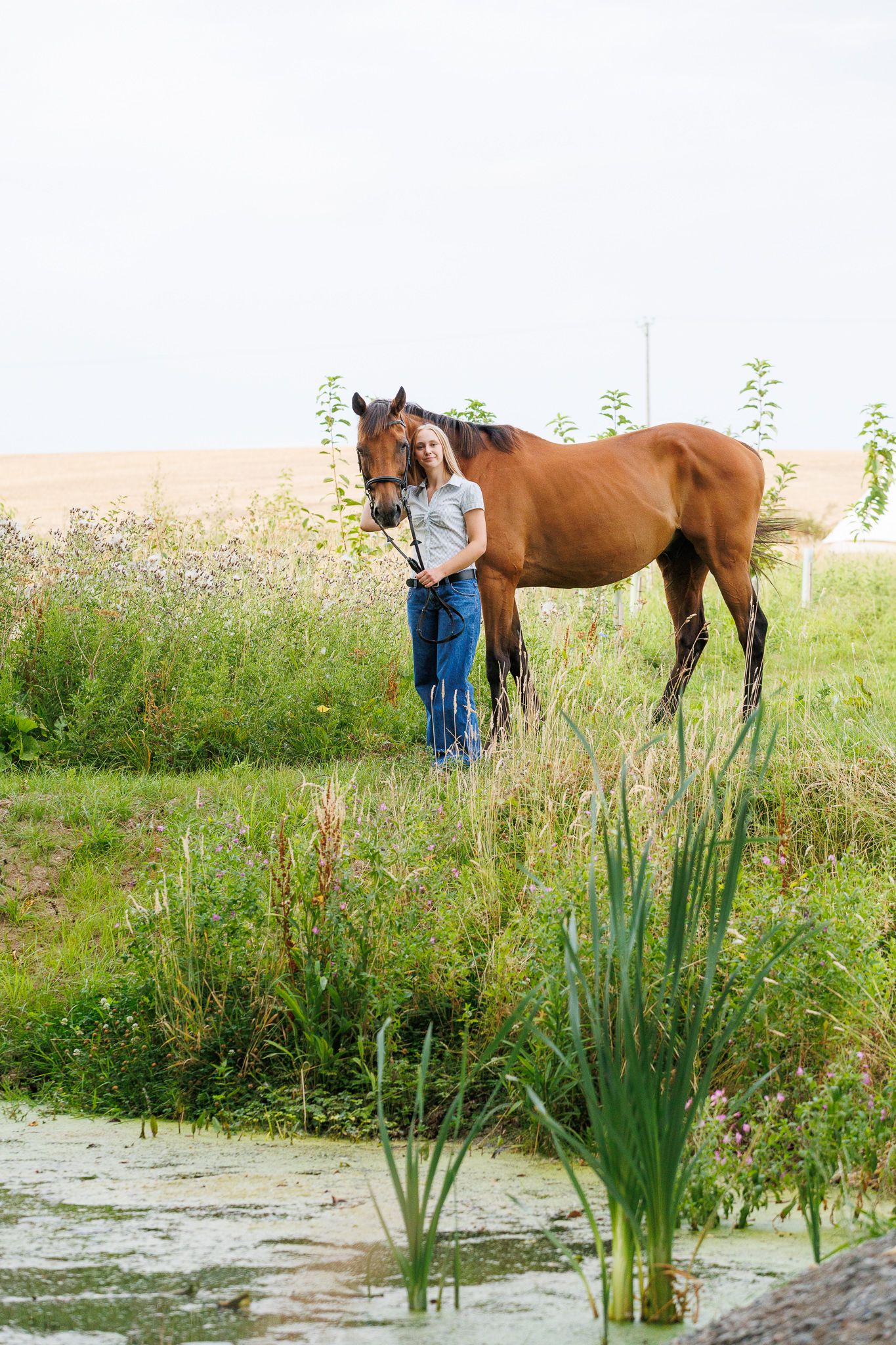 Young woman smiling between two bay horses with white markings, holding reins near a brick stable.