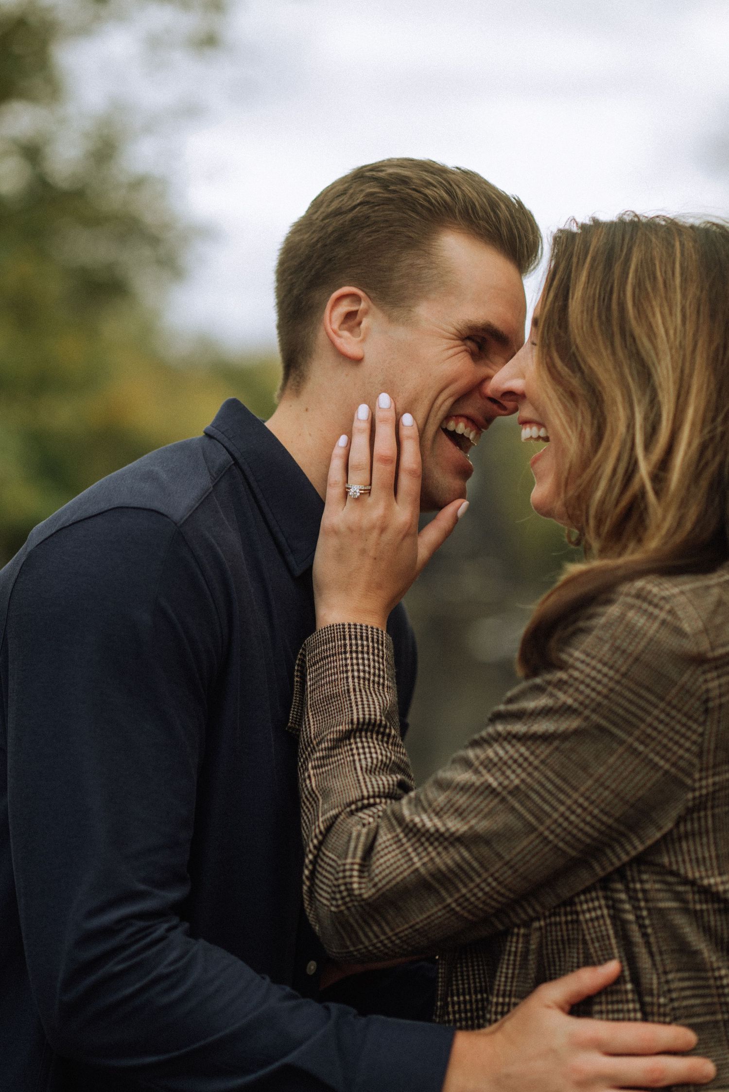 Smiling couple share an intimate moment outdoors as she cups his cheek, showing off a sparkling engagement ring.