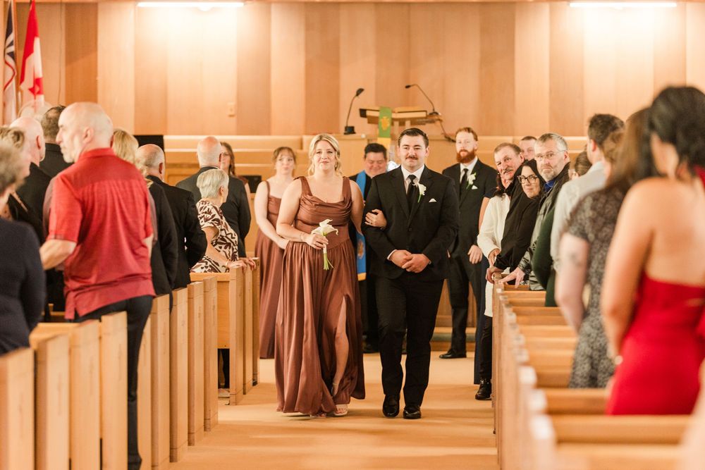 Groomsman escorts bridesmaid down church aisle during wedding ceremony with guests standing in wooden pews.
