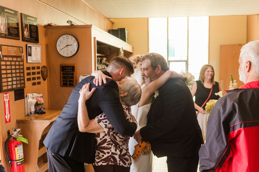 Wedding guests share a warm group hug in a decorated indoor venue with a clock on the wall.