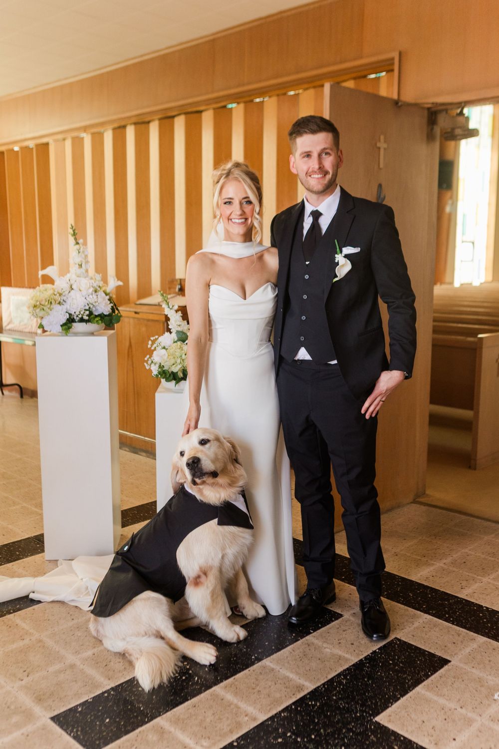 Bride and groom posing with their golden retriever dressed in a tuxedo inside a church on their wedding day.