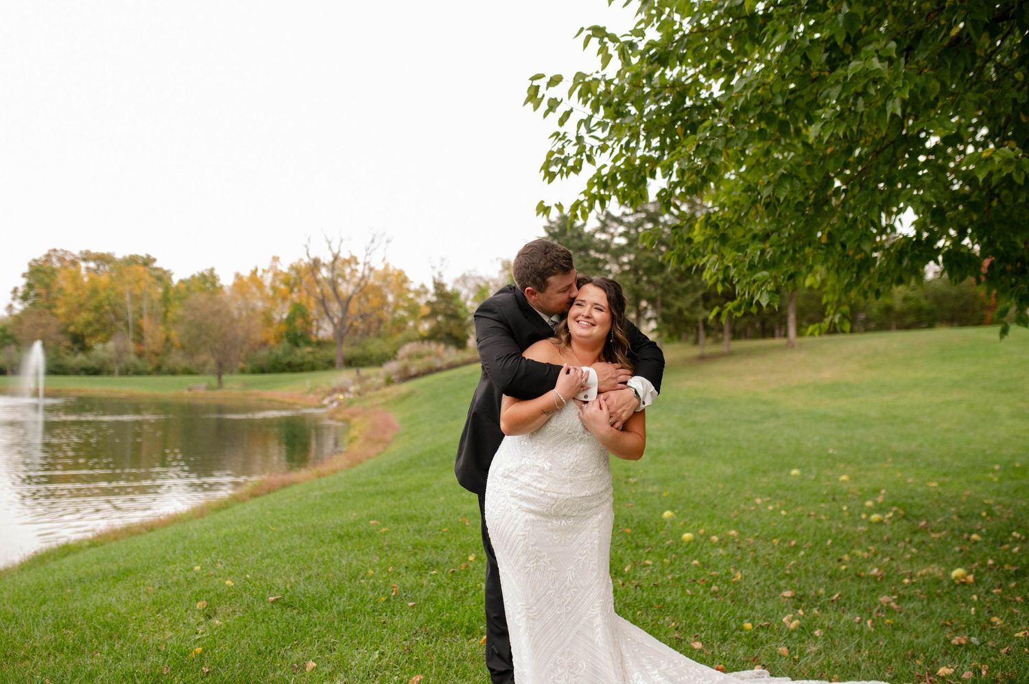 Bride and groom embrace outdoors near a pond with autumn trees in background at wedding.