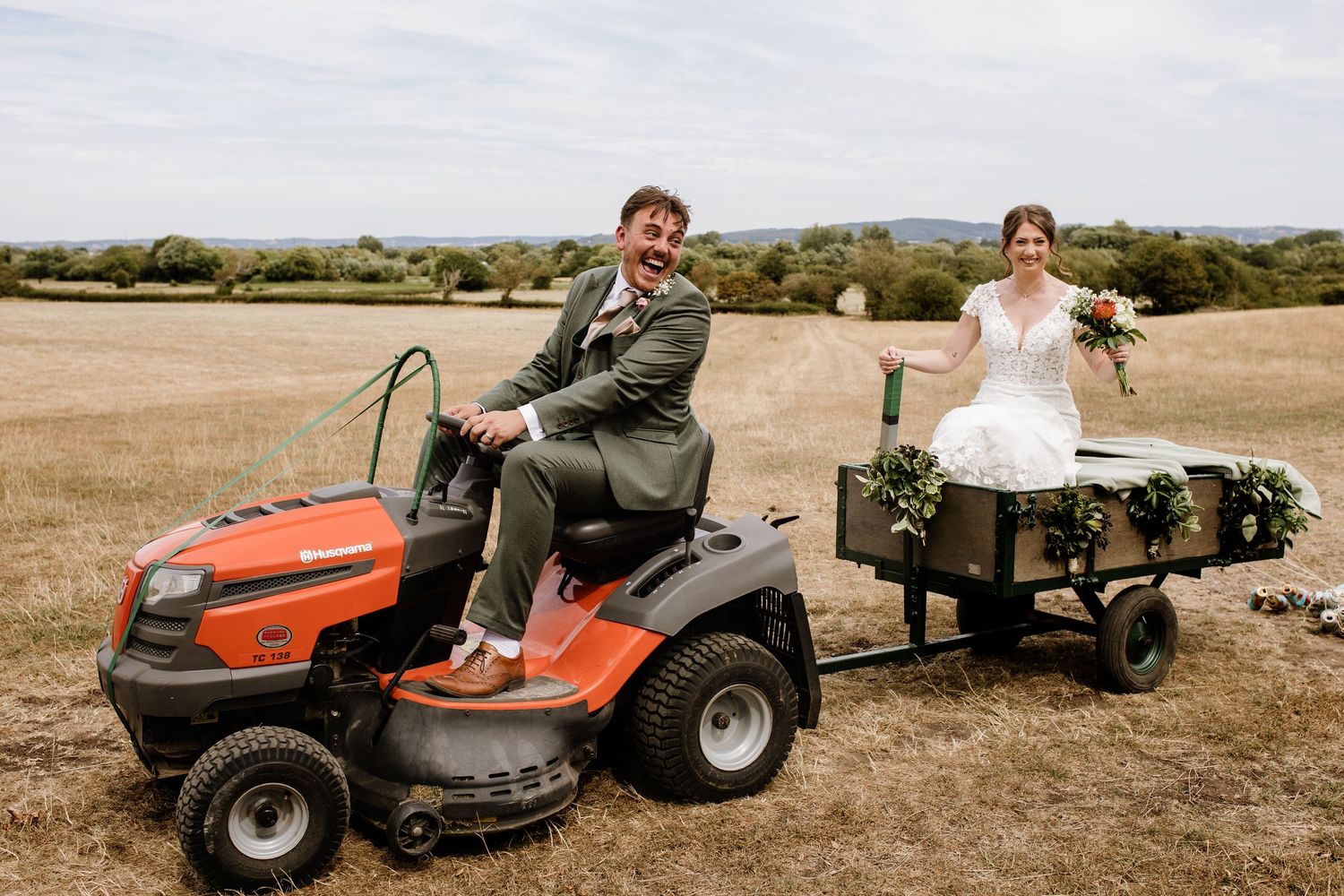 Groom rides orange lawn tractor pulling bride in decorated cart across rustic field at outdoor wedding.