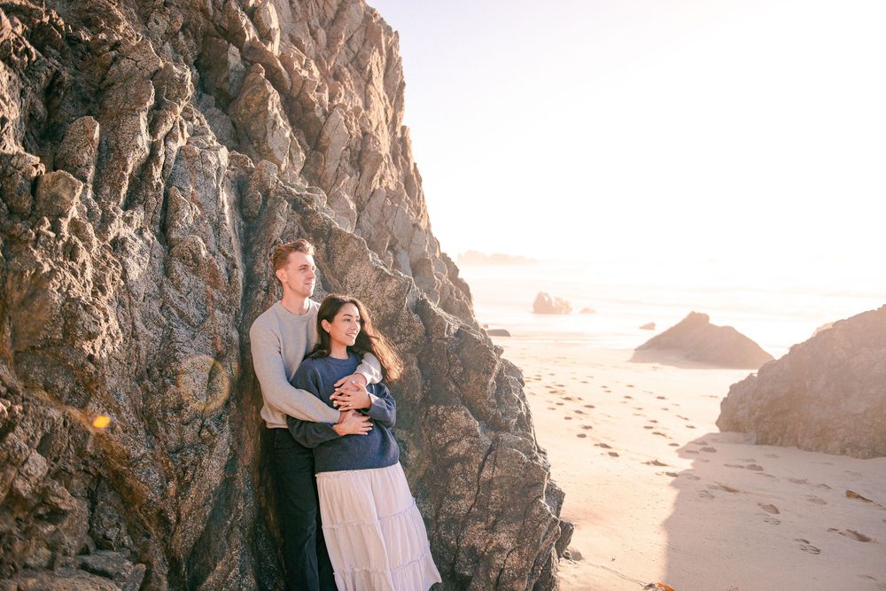 Couple embracing against rocky cliffs on a sunlit beach, with ocean waves and golden light in the background.