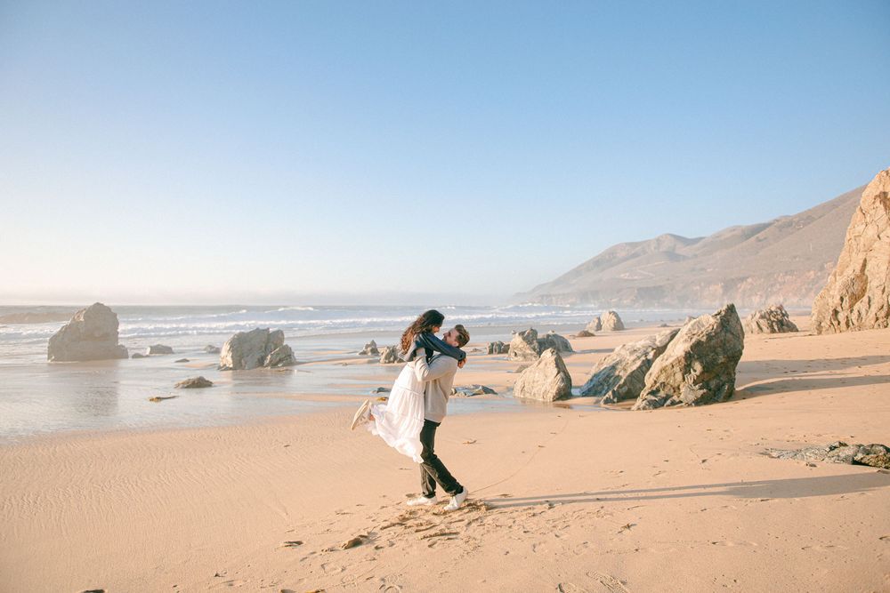 Couple embracing on a sandy beach with rocky coastline and clear blue sky in the background.