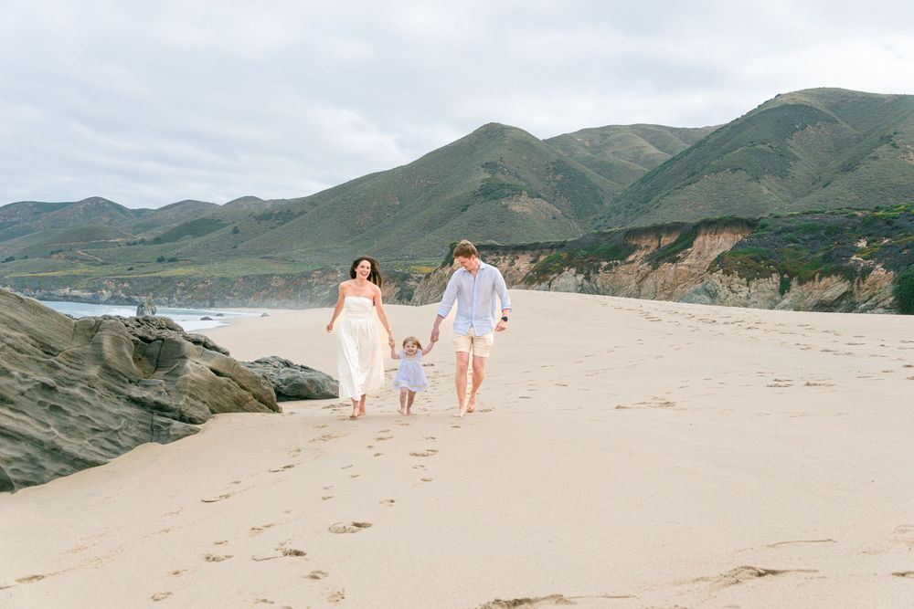Family of three walking hand-in-hand on a sandy beach with green mountains and rocky cliffs in the background.