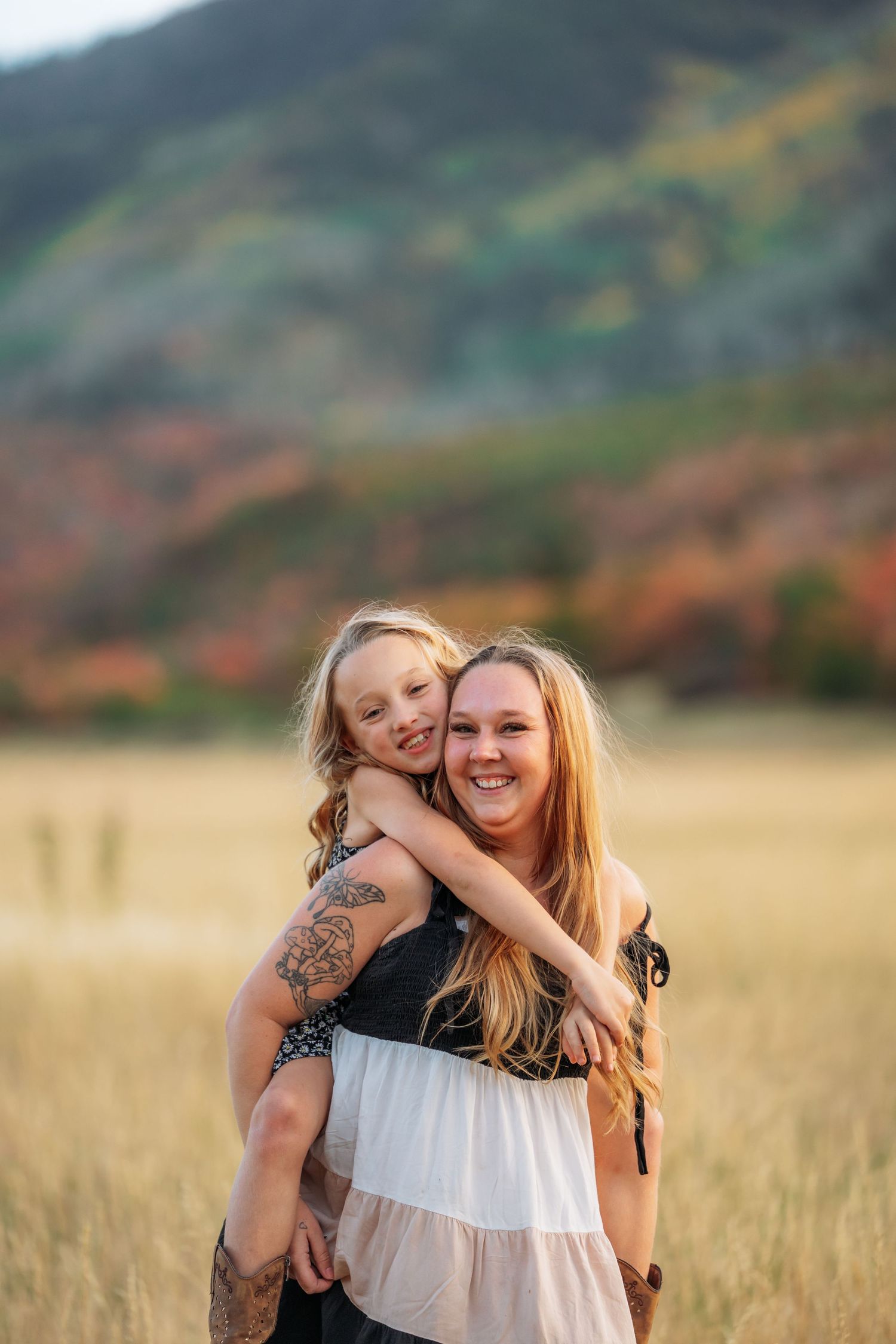 A woman with a tattoo smiles as a young girl hugs her from behind in a golden field with autumn hills.