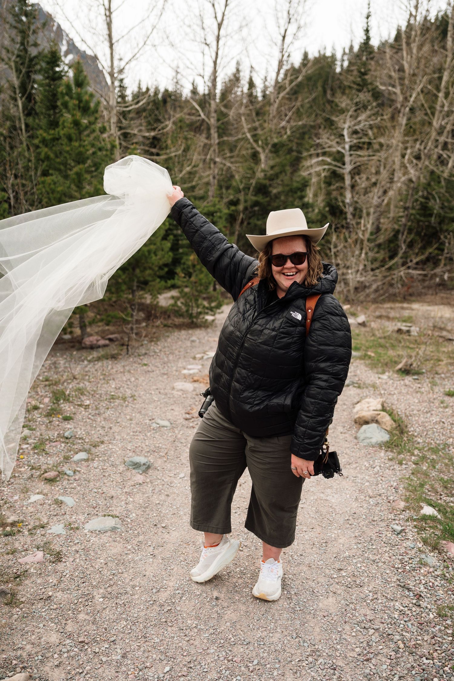 Woman in cowboy hat holds flowing veil on mountain trail surrounded by pine trees and rocky landscape.