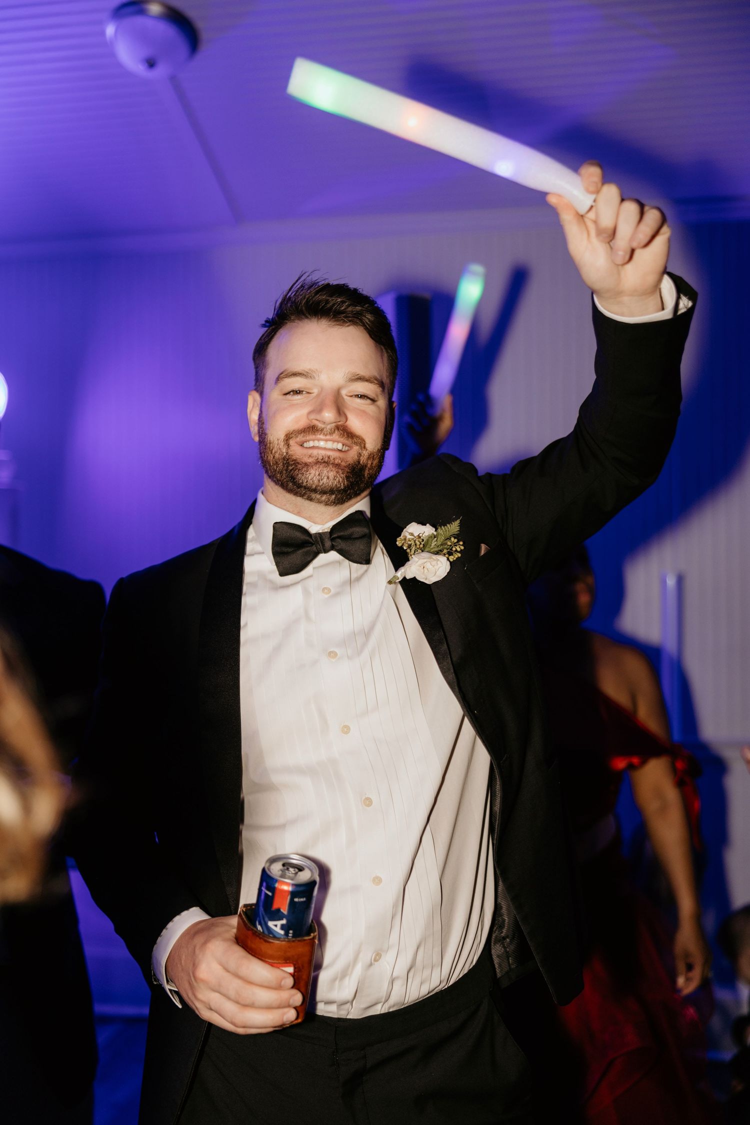 Smiling groom in black tuxedo holds beer can and raises glow stick on wedding reception dance floor with purple lighting.