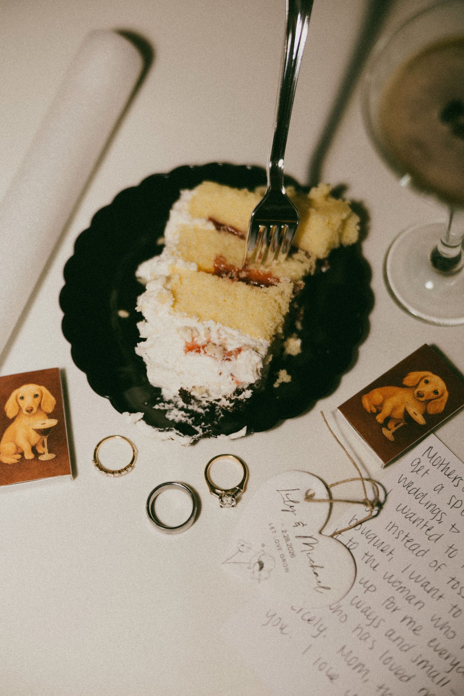 Wedding cake slice on black plate with gold rings, handwritten note, dachshund cards, and wine glass on white table.