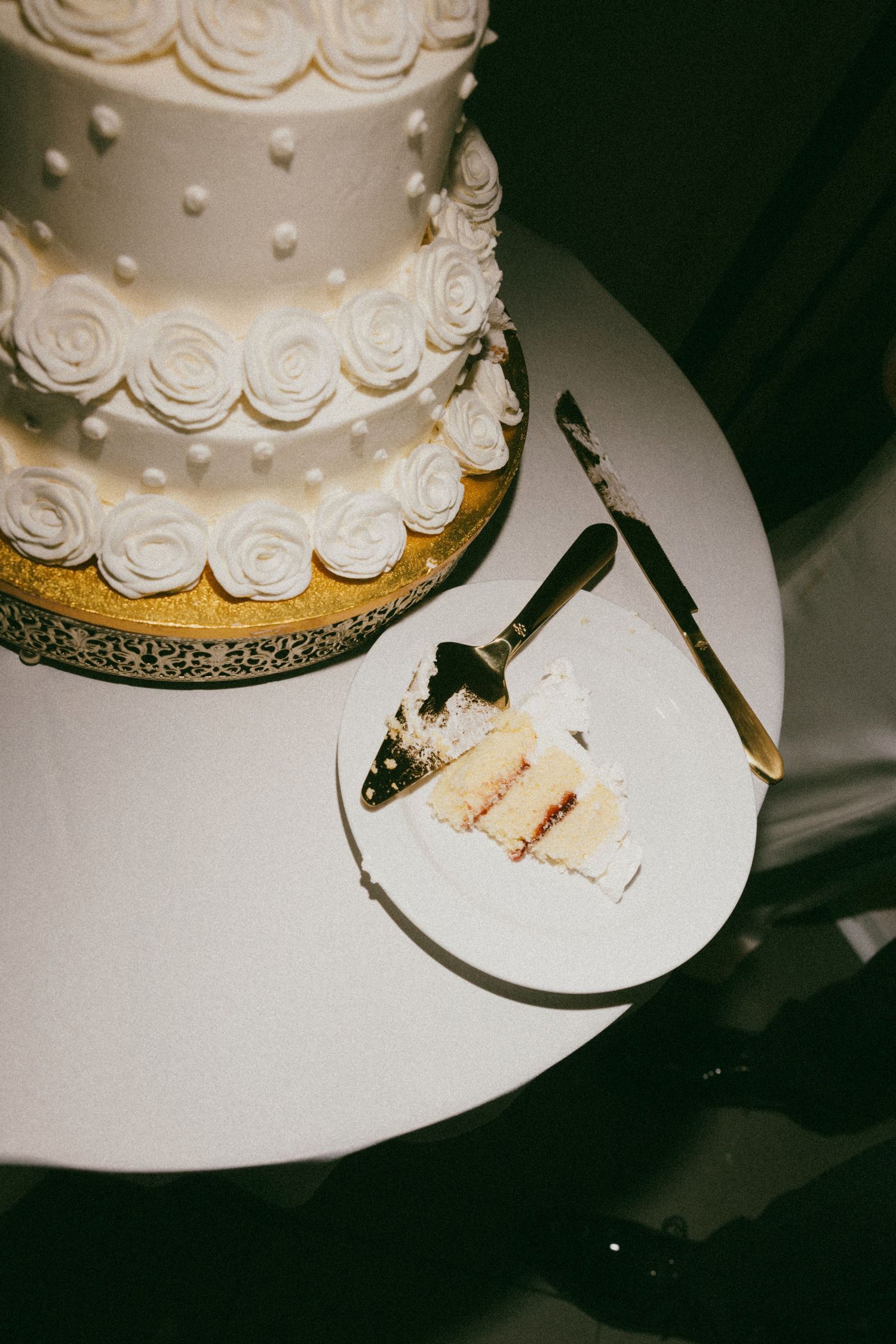 Elegant white wedding cake with rose details beside a plated slice and gold cake knife on a round table.