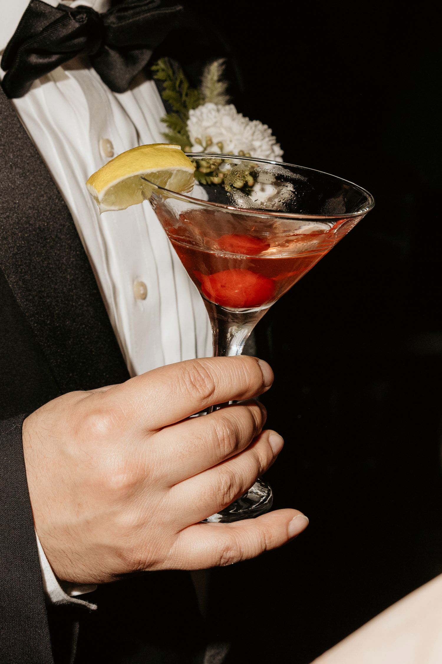 Groom in tuxedo holding a red martini cocktail garnished with lemon slice and cherries at a wedding reception.