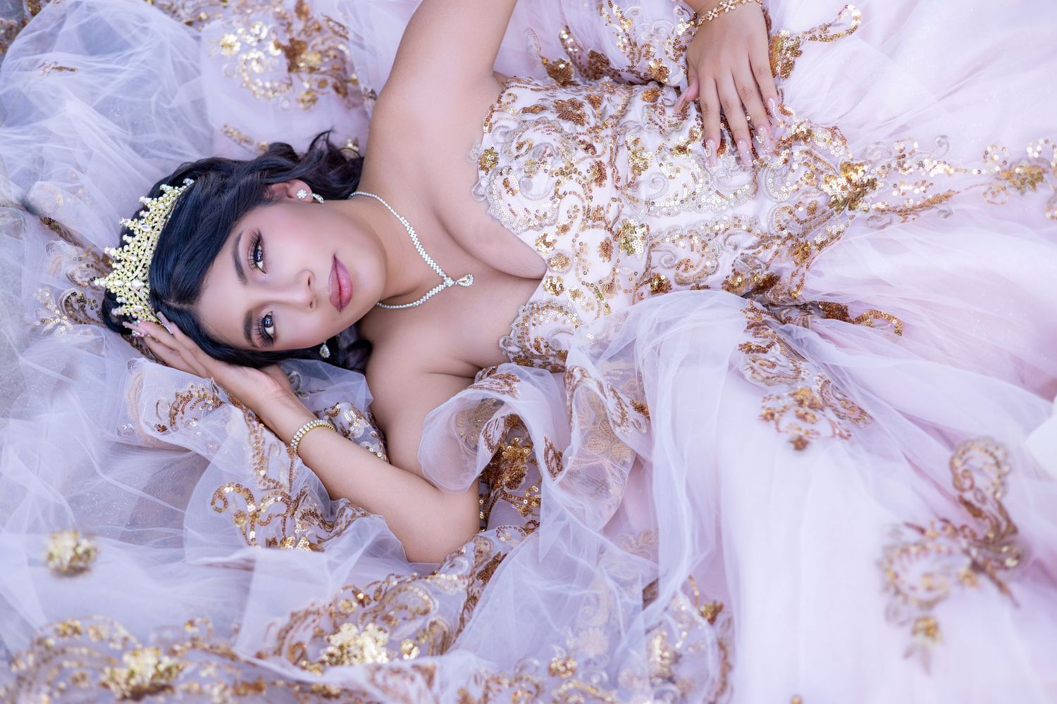 Young woman in elegant white and gold ball gown with tiara lying gracefully among flowing tulle fabric.