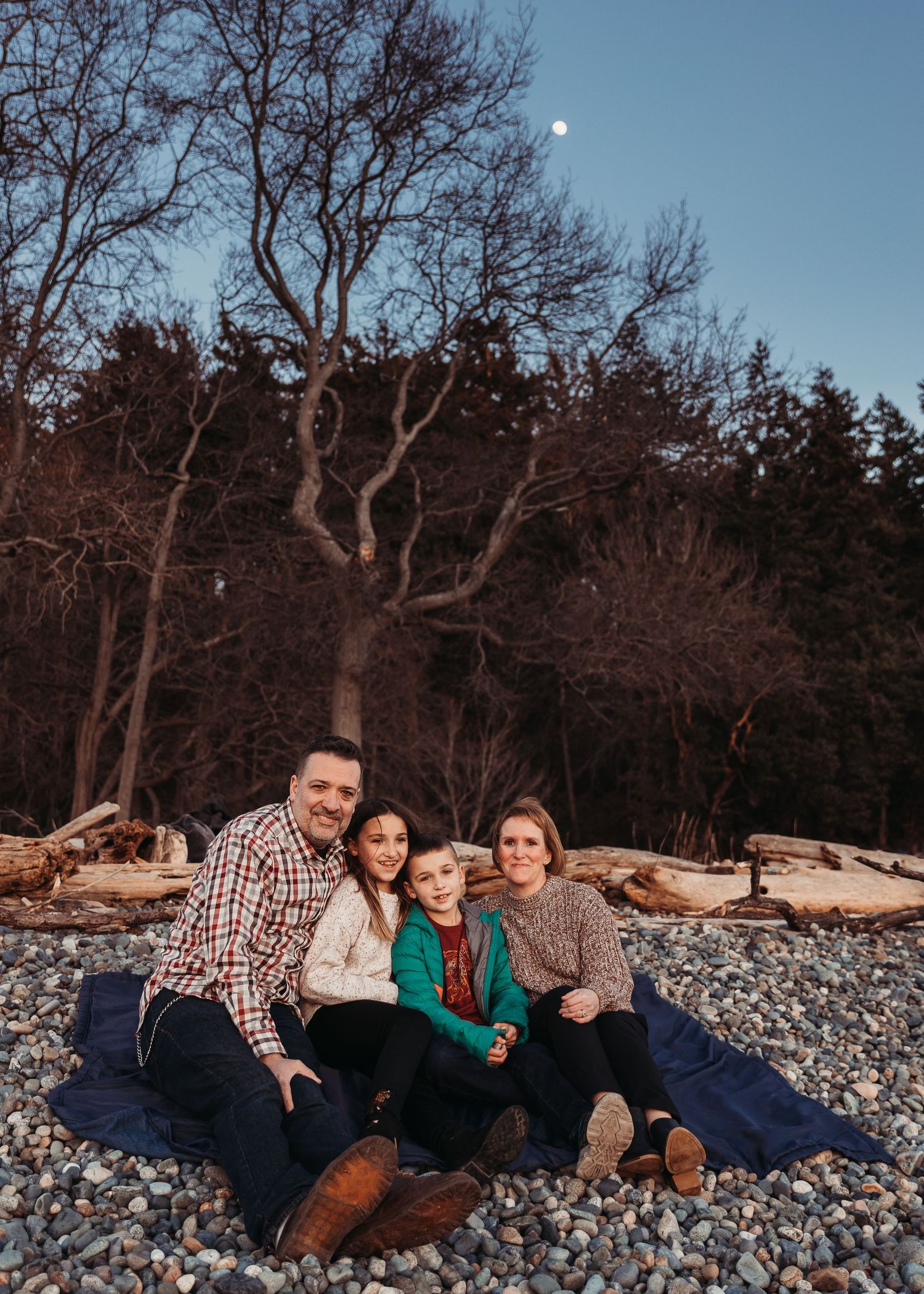 Family of four sitting on a blue blanket on a pebble beach at dusk with driftwood, bare trees and a moon in the sky.