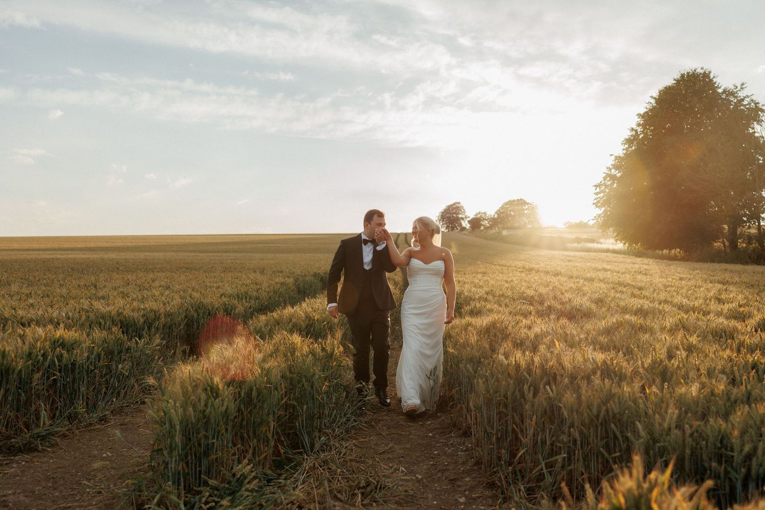 Bride in a white gown and groom in a dark suit walk hand in hand along a path through a golden wheat field at sunset.