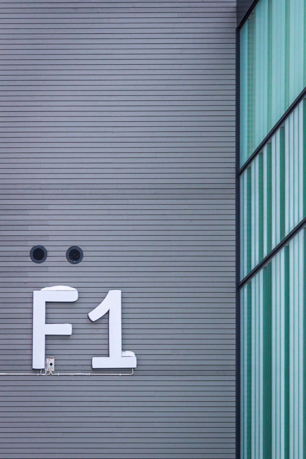 White F1 sign on gray corrugated metal wall beside teal green striped building facade.