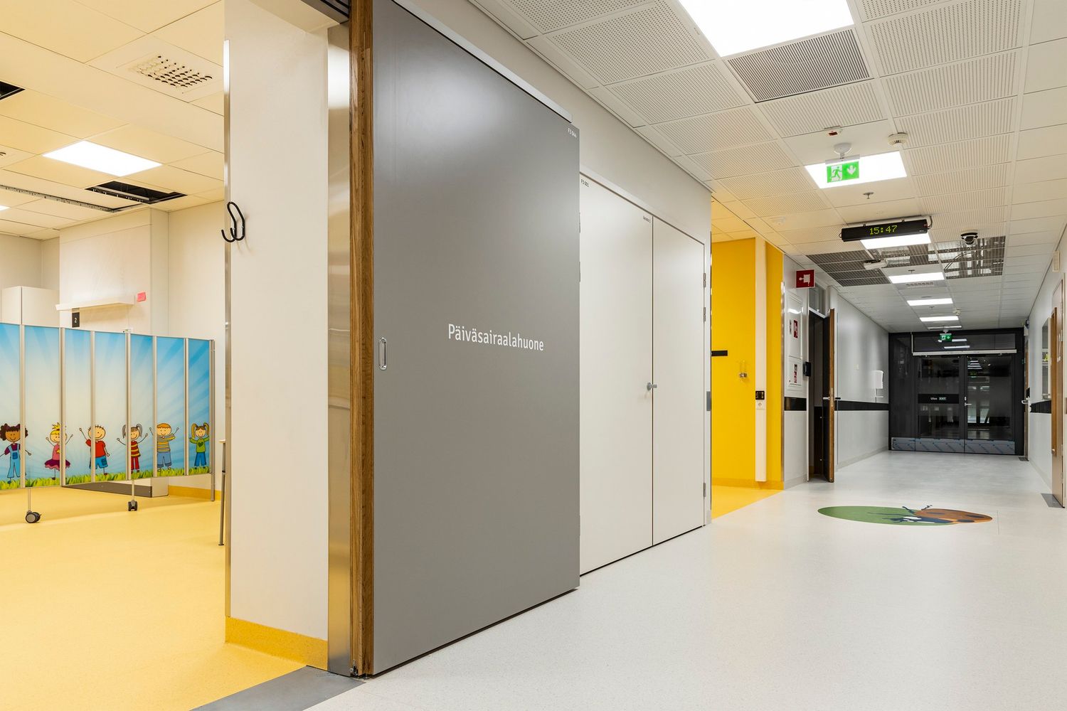 Modern hospital corridor with yellow accents, gray sliding door, and colorful wall murals in a pediatric ward.
