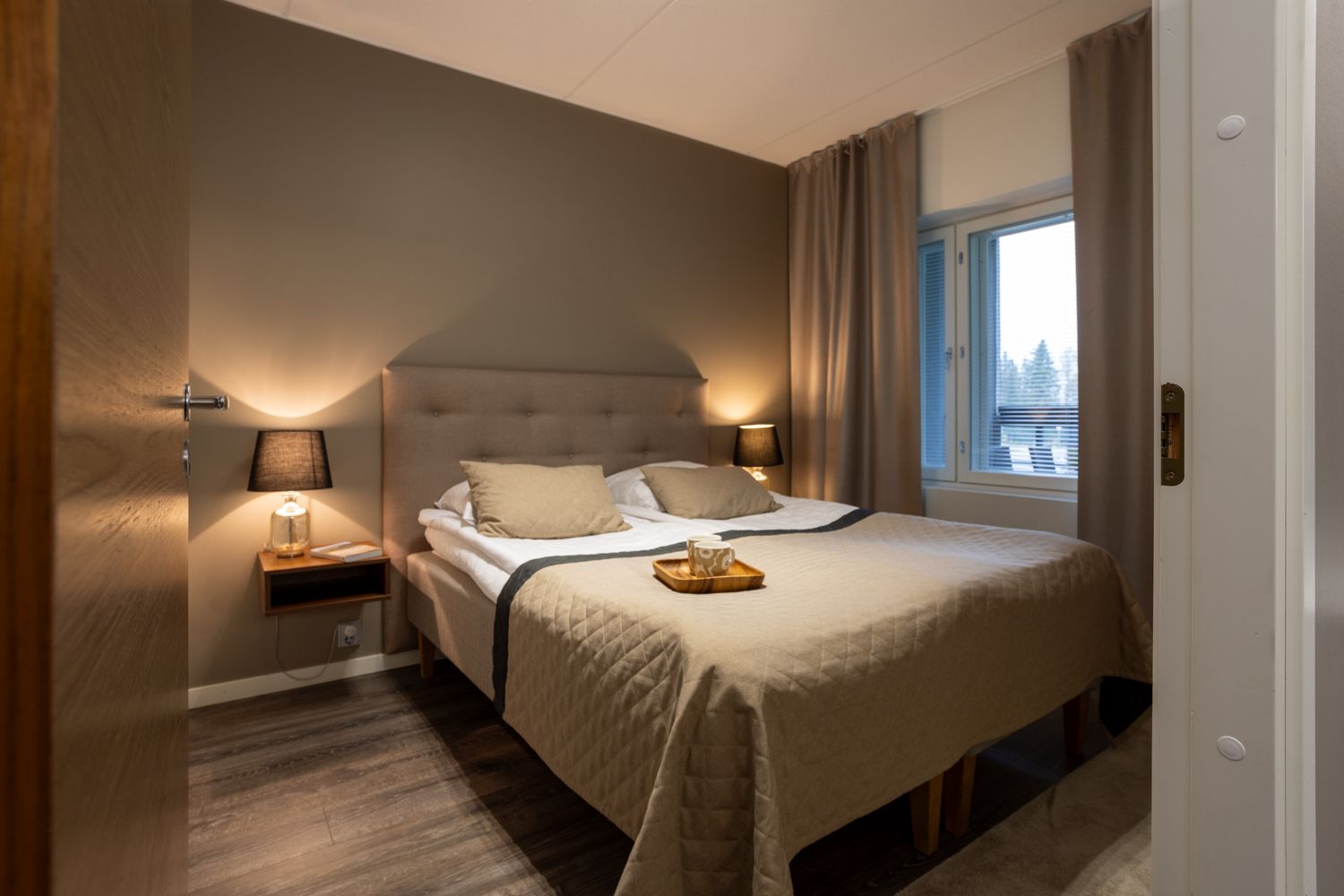 Cozy hotel bedroom with a double bed, warm lighting, neutral tones, wooden floors, and a window with curtains.