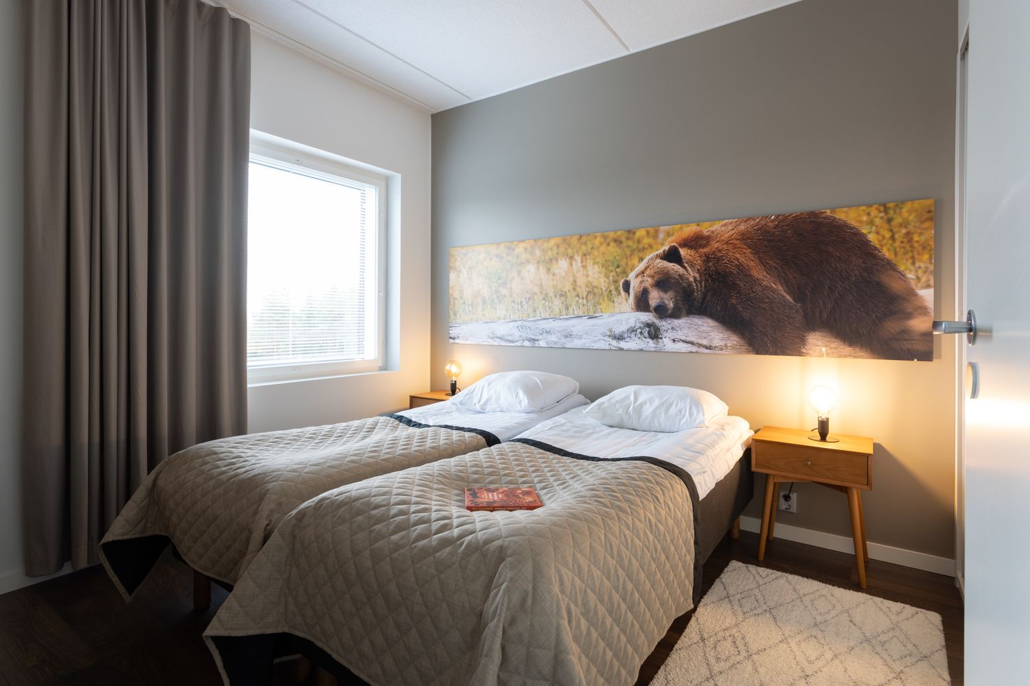 Cozy hotel twin bedroom with bear artwork above bed, warm lighting, neutral tones, and a window with curtains.