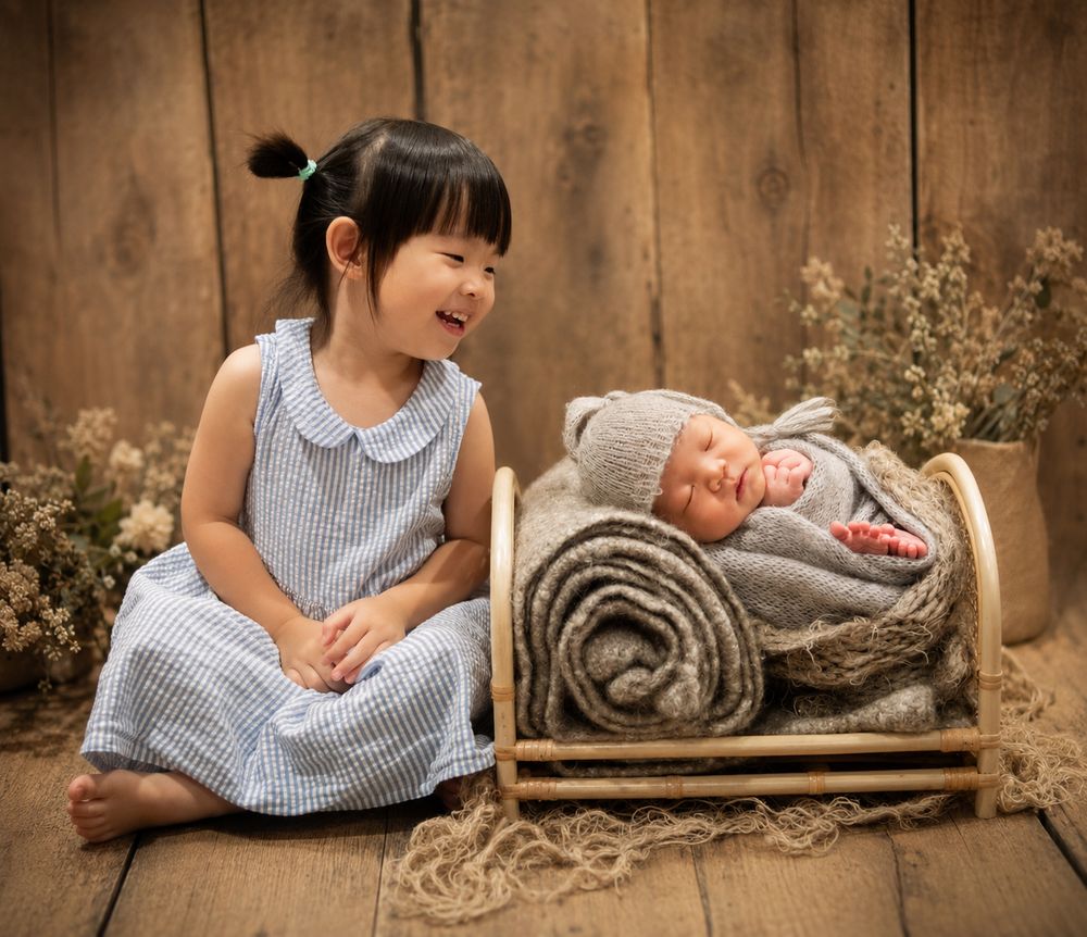 A smiling toddler girl in a blue dress sits beside a sleeping newborn baby wrapped in gray knit on a wooden prop bed.