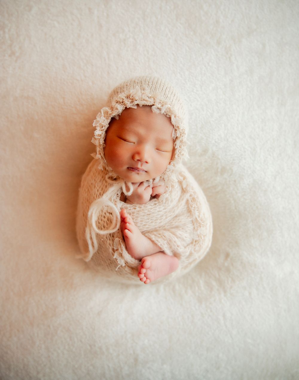 Sleeping newborn baby wrapped in cream knit blanket and bonnet with floral trim on white fluffy backdrop.
