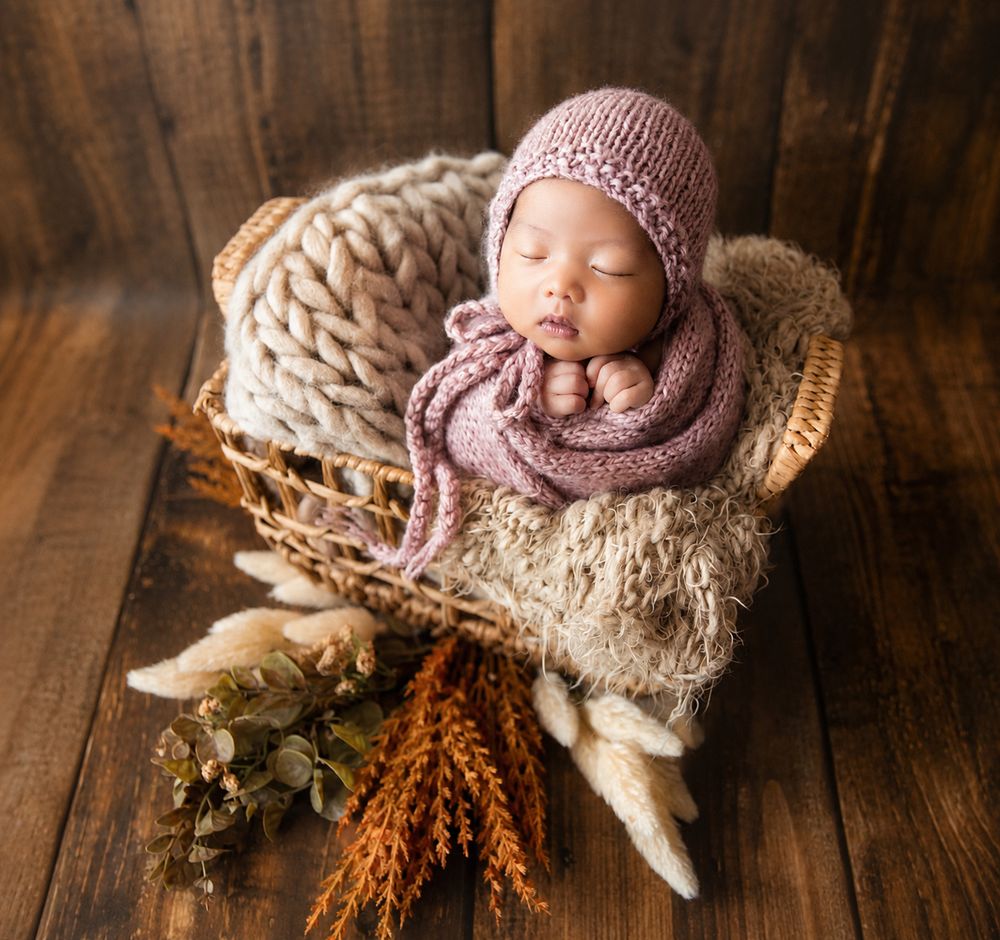 Newborn baby sleeping peacefully in a wicker basket, wrapped in a pink knit blanket and bonnet, surrounded by dried flowers.