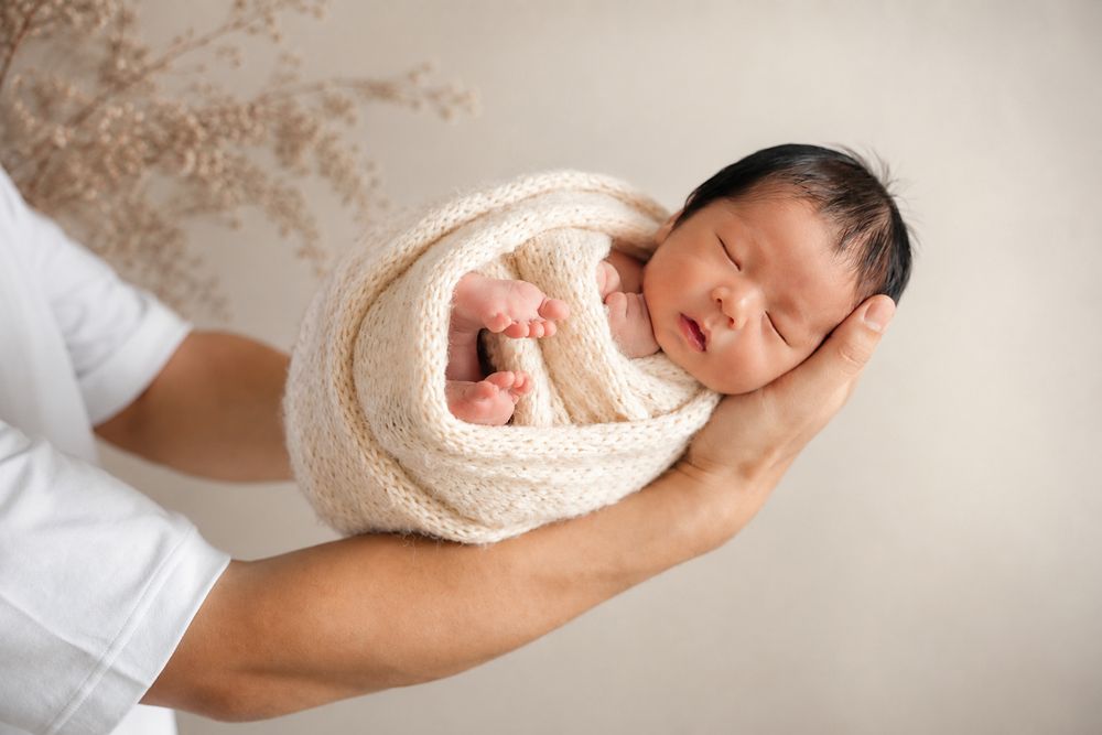 A newborn baby swaddled in cream wrap sleeping peacefully on an adult hand in a soft neutral studio setting.
