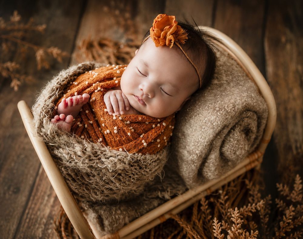 Newborn baby sleeping peacefully in a tiny wooden bed, wrapped in orange knit blanket with floral headband.