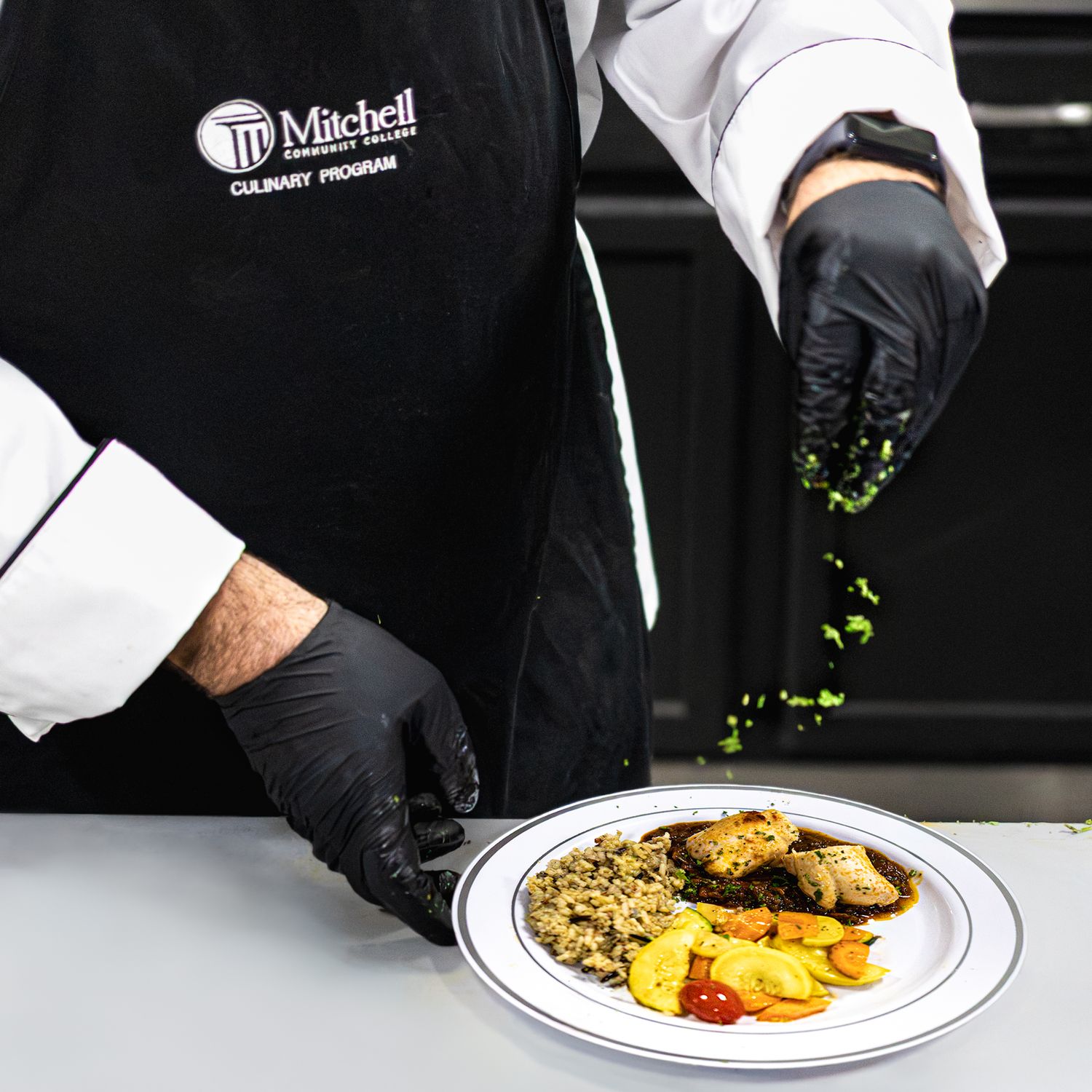 Chef in Mitchell Culinary Program apron garnishing a plated dish with grains, roasted vegetables, and protein.