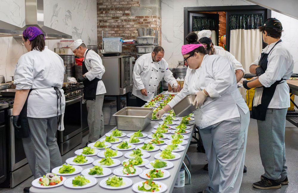 A culinary team plates green salads on a long row of white dishes in a professional kitchen.