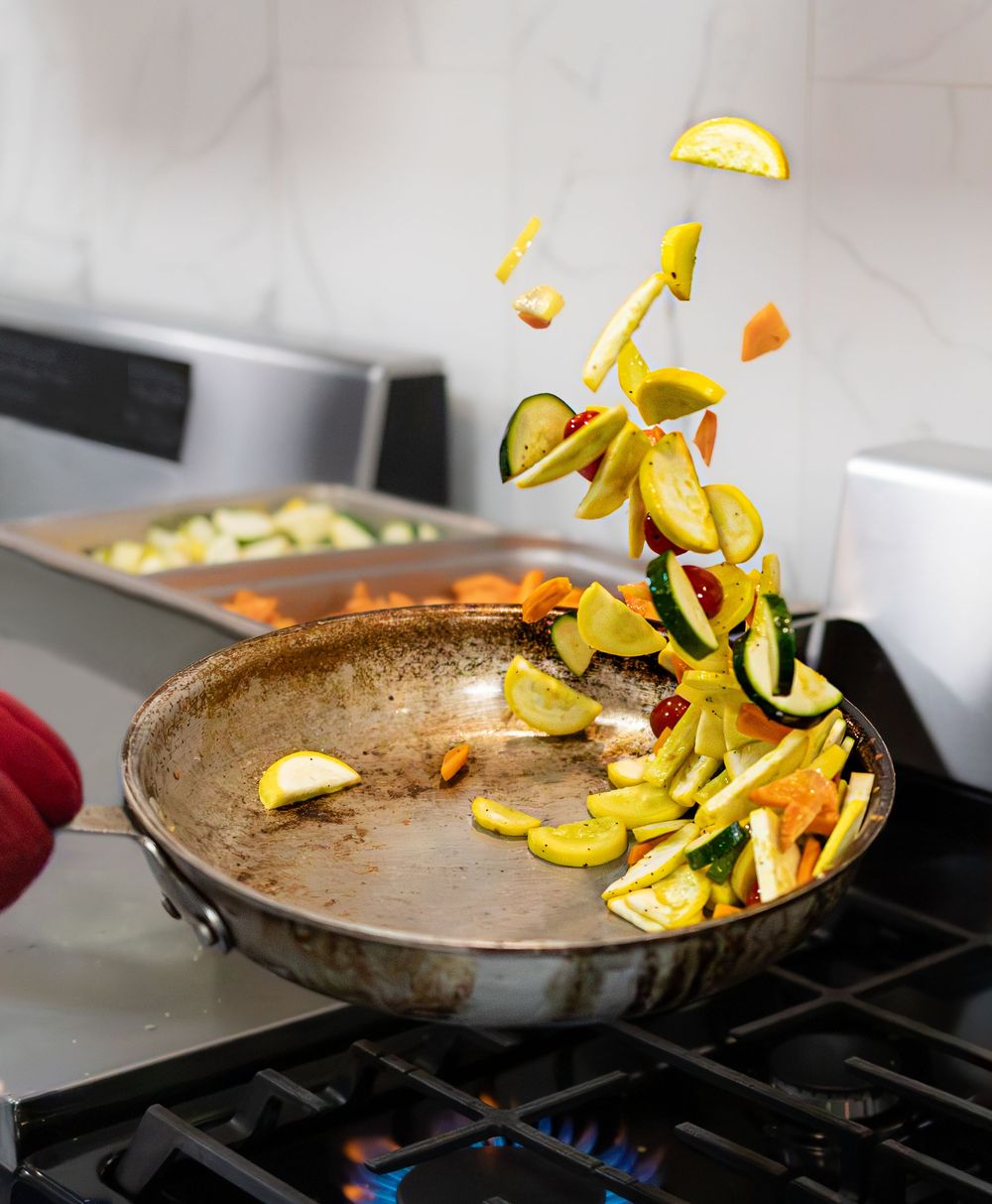 Colorful sliced vegetables being tossed in a well-seasoned pan on a gas stove.