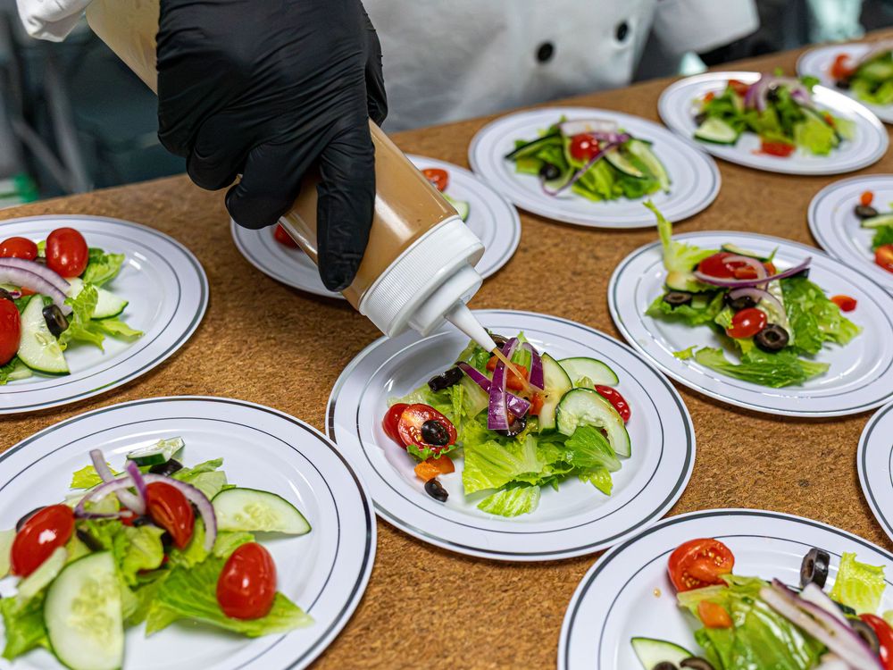 A gloved hand drizzles dressing over multiple fresh garden salads on white plates.