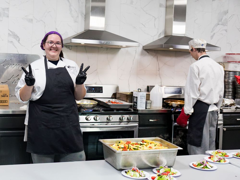 A cheerful chef poses with peace signs in a professional kitchen with plated dishes on the counter.
