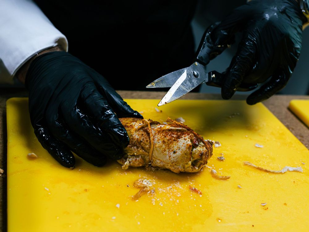 Gloved hands use scissors to cut a roasted stuffed meat roll on a yellow cutting board.