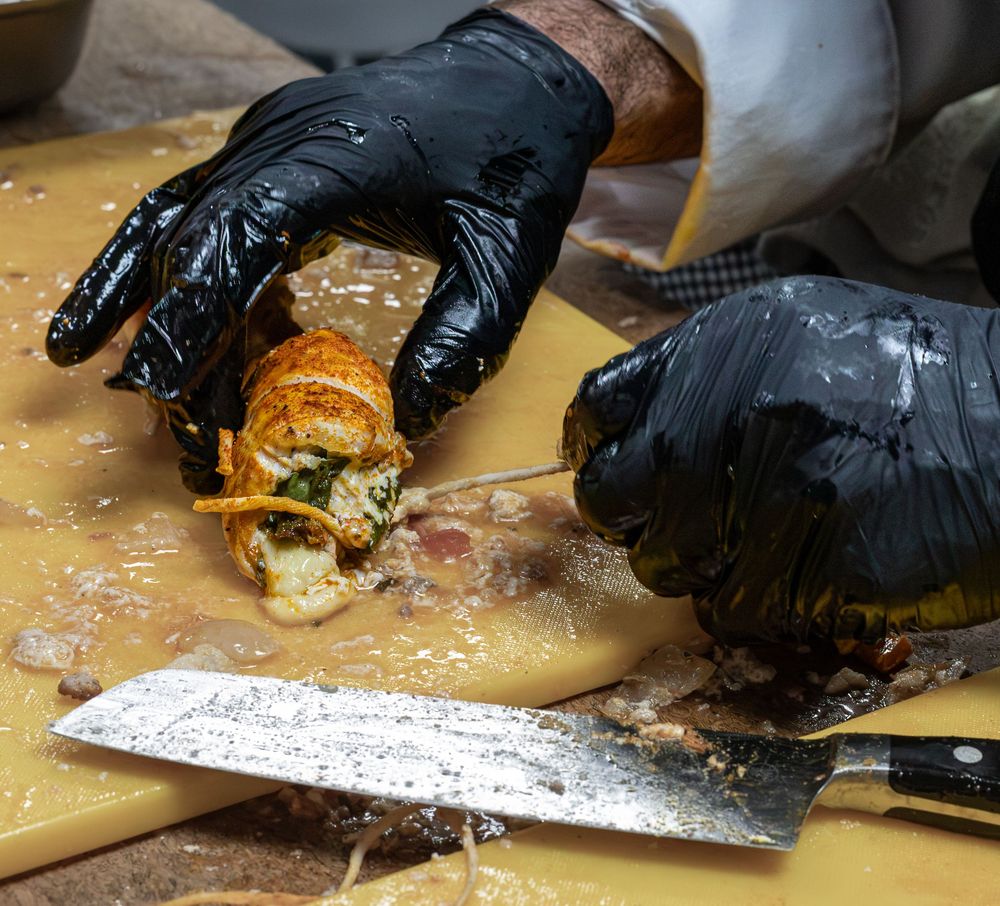 A gloved chef slices a bacon-wrapped stuffed meat roll on a yellow cutting board with a knife.
