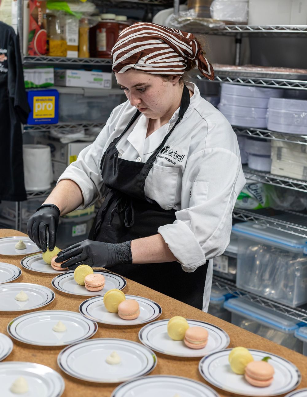 A culinary student carefully plates colorful macarons on white dishes in a professional kitchen.