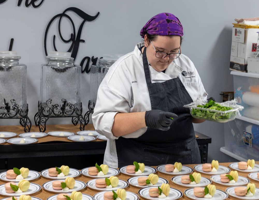 A chef in a purple headscarf garnishes elegantly plated dessert dishes at a catering event.