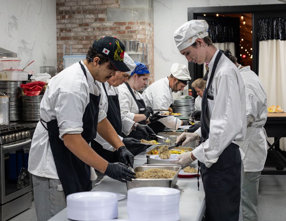 A team of culinary students in chef whites prepare and plate food in a busy restaurant kitchen.