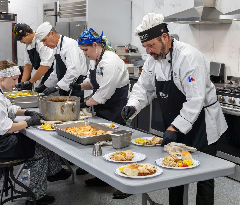 Culinary students in chef whites plate food together at a stainless steel table in a cooking class.