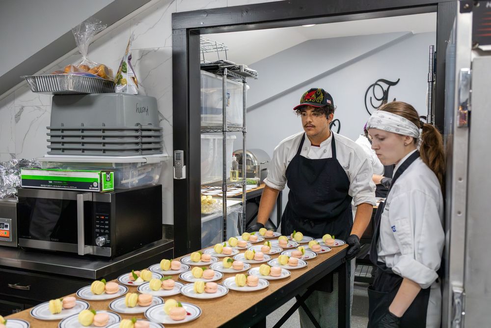 Two culinary students review plated desserts on a prep counter in a professional teaching kitchen.