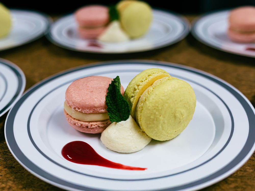 Two colorful French macarons with cream and raspberry sauce on a white plate.