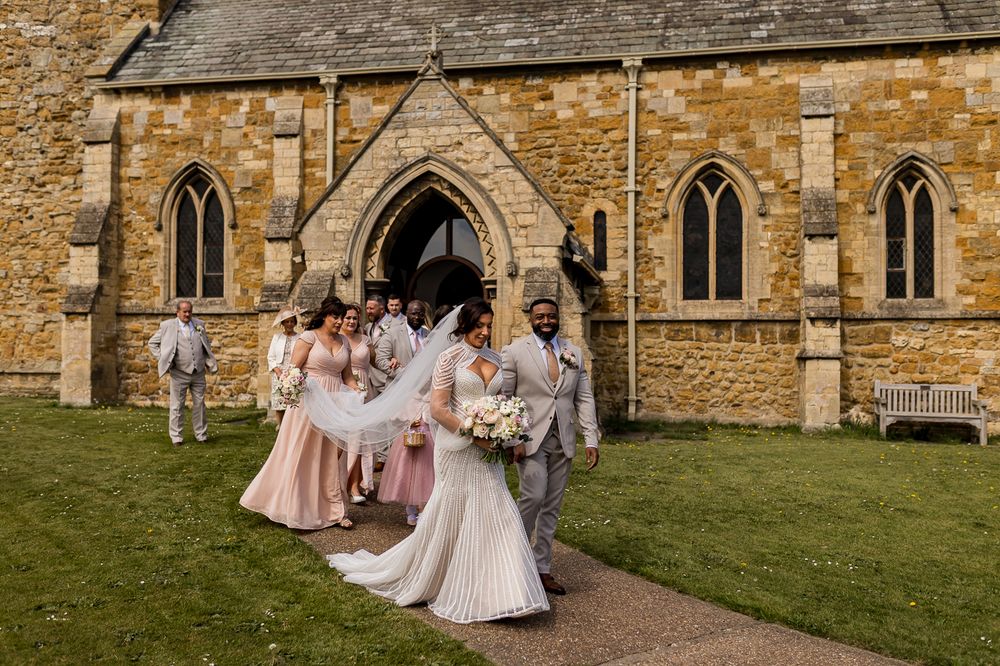 Wedding party exits stone church, bride in white gown with bridesmaids in pink dresses and groomsmen in gray suits.