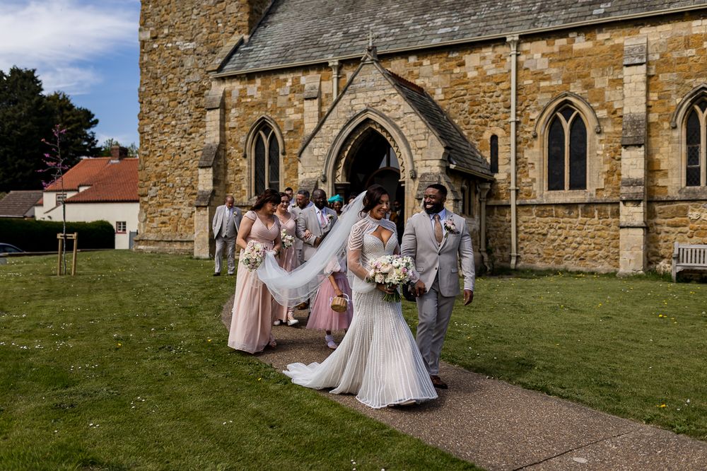 Bride and groom exit stone church with bridesmaids in pink dresses and groomsmen in grey suits on wedding day.