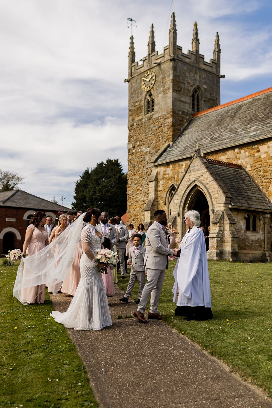 Wedding party outside a historic stone church, bride in white gown with veil, greeted by officiant on sunny day.