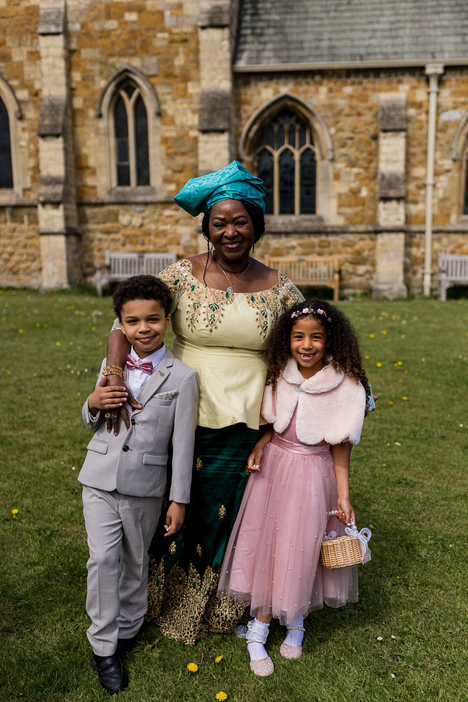Woman in traditional African attire poses with two children in formal wear outside a stone church.