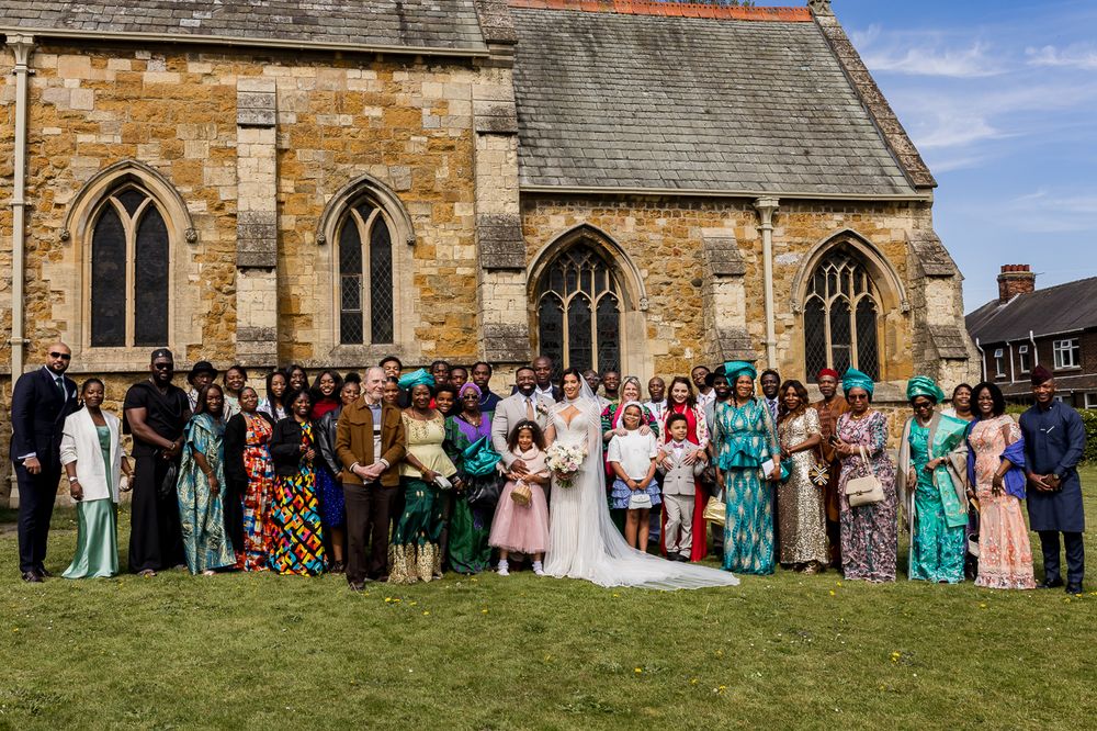 Wedding party posing outside a stone church, featuring bride in white gown and guests in colorful traditional African attire.