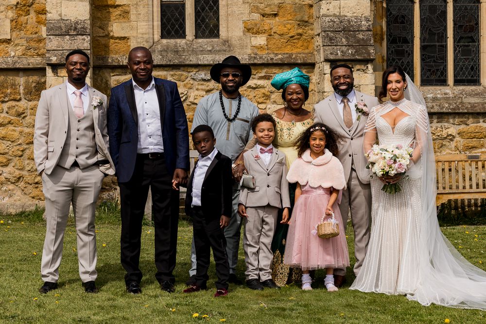 A joyful wedding party poses outside a stone church, featuring the bride in white, groom, family members, and children in formal attire.