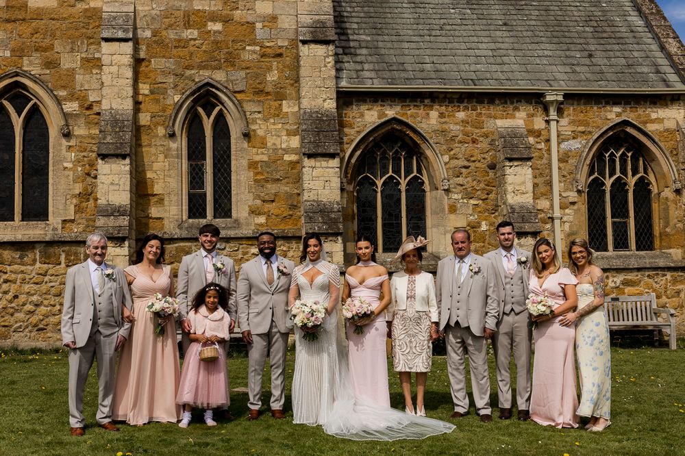 Wedding party posing outside a historic stone church on a sunny day in elegant attire.